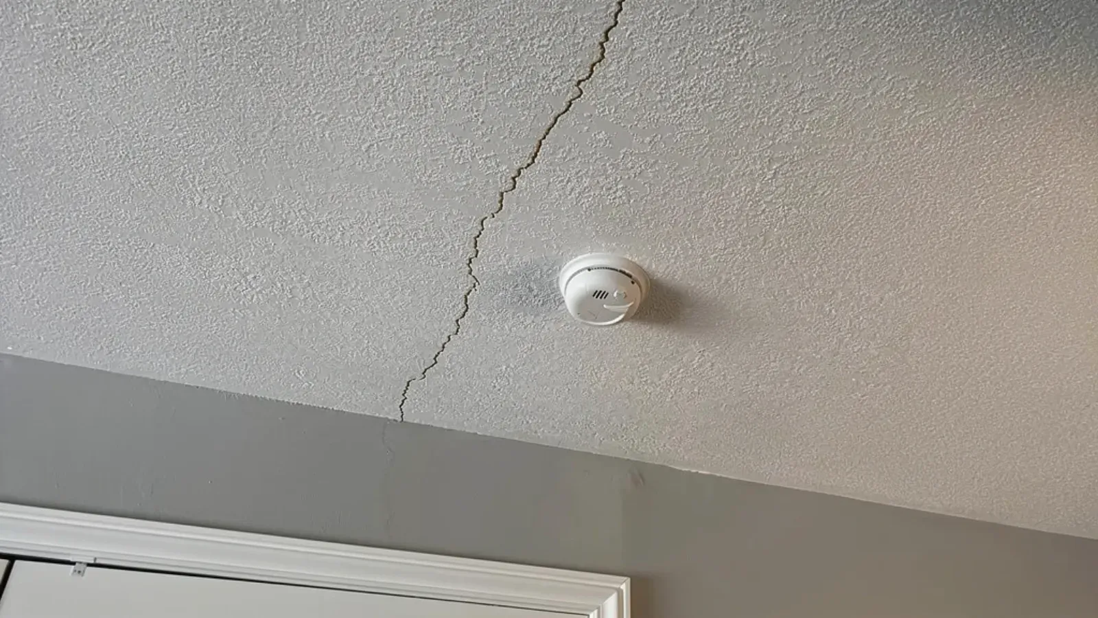 A fine hairline crack running across a lightly textured painted ceiling toward the corner where the ceiling meets the wall, with a round white smoke detector mounted nearby