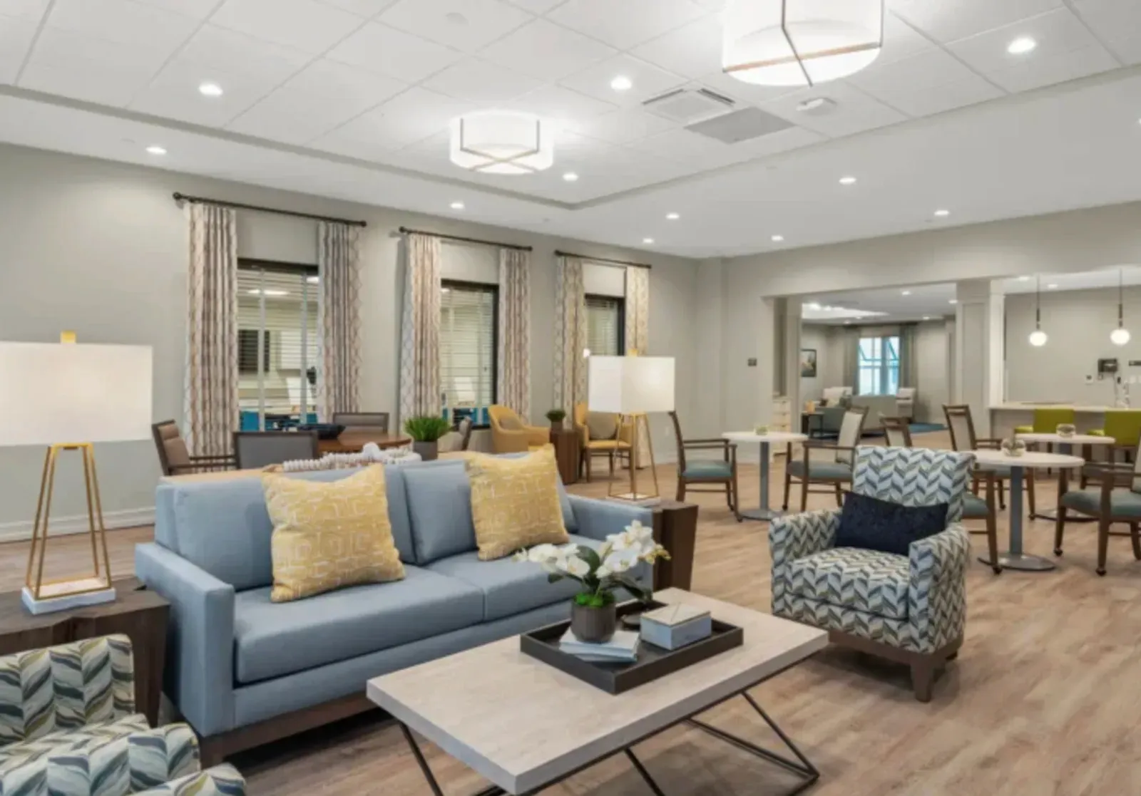 Senior-living community lounge with finished painted drywall walls, a suspended ceiling with recessed and pendant lighting, curtained windows, and upholstered sofas and armchairs around a coffee table